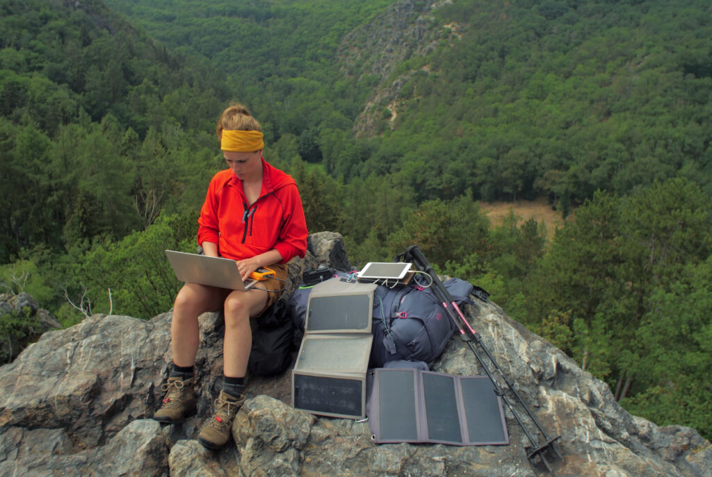 Frau mit braunen Wanderschuhen, roter Jacke und gelbem Stirnband sitzt mit Laptop auf den Knien auf der Bergspitze, rechts daneben Rucksack, Wanderstöcke und faltbare Solarmodule 