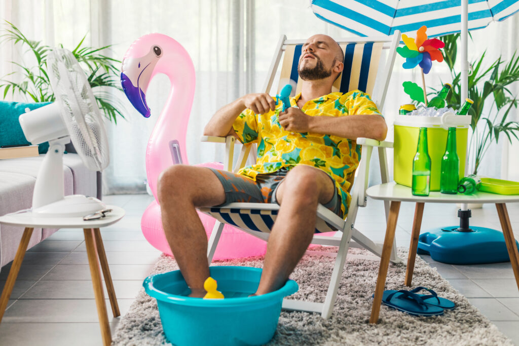 Mann in Shorts und Hawaihemd sitze im Raum im Liegestuhl, die Füße in einer Schüssel mit Wasser. In dieser schwimmt ein Quietscheentchen. Im Hintergrund Teil eines gestreiften Sonnenschirms sowie eines Plastikflamingos