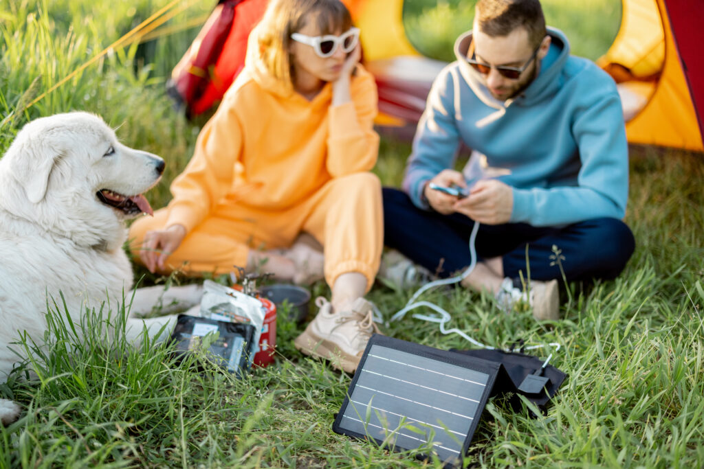 Eine Frau,links, und ein Mann, rechts sitzen vor einem Zelt. Beide haben Sonnenbrillen auf. Er hat ein Handy in der Hand, welches an ein mobiles Solarmodul, etwa in Ipad-Größe, das vor ihm auf dem Rasen steht, angeschlossen ist